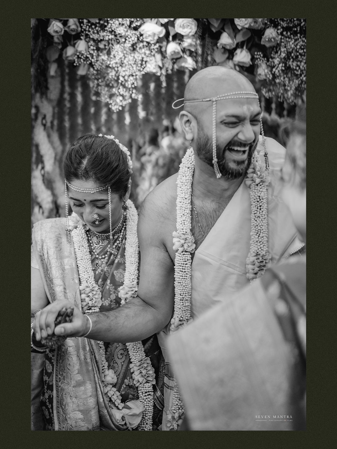 Black and white photo of laughing Indian bride and groom in flower garlands during wedding rituals.