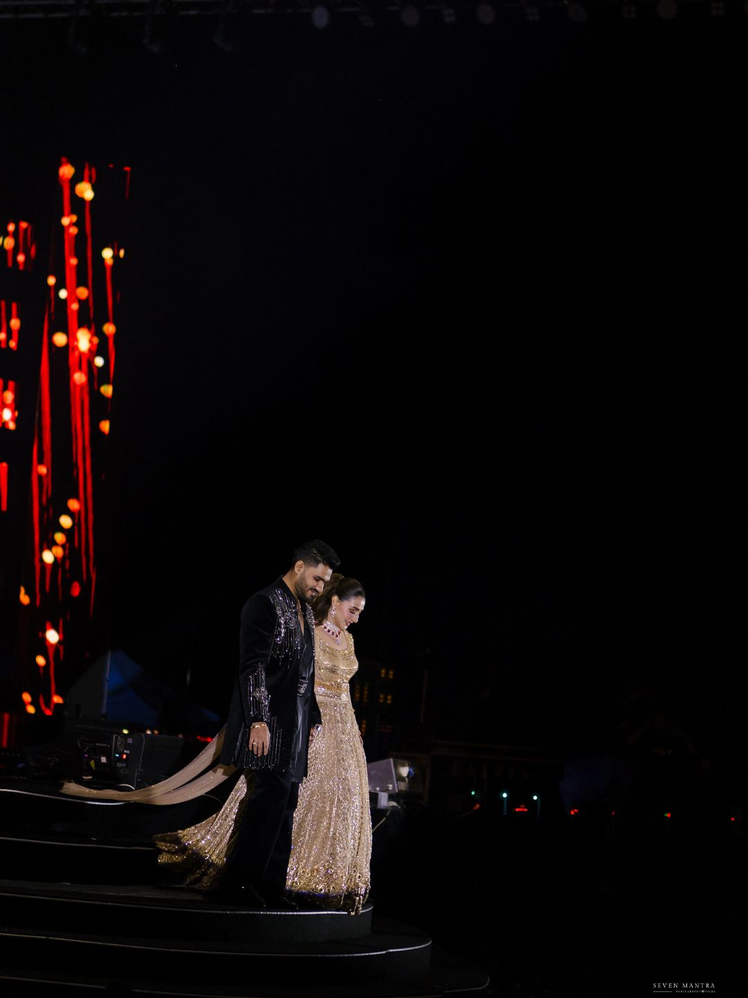 Bride and groom silhouetted on stage with dramatic red hanging lights at Indian wedding.