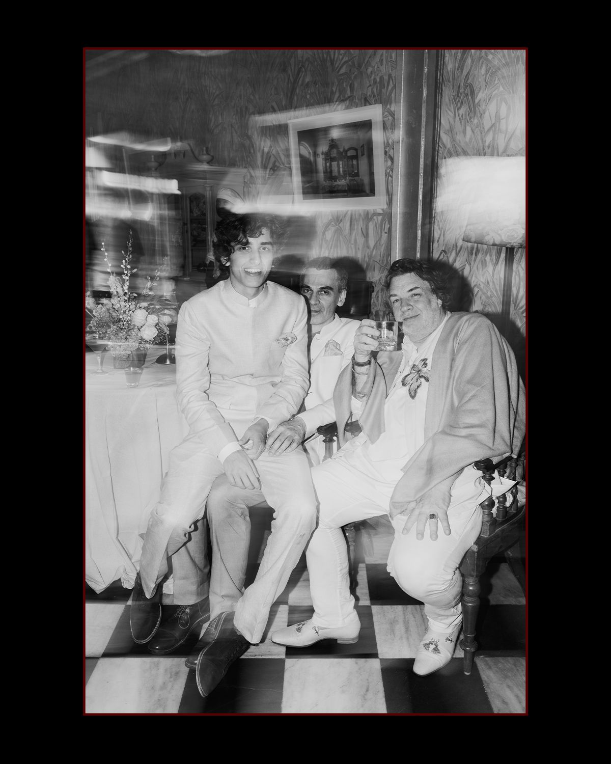 Black and white photo of three guests sitting together at an Indian wedding reception on checkered floor.