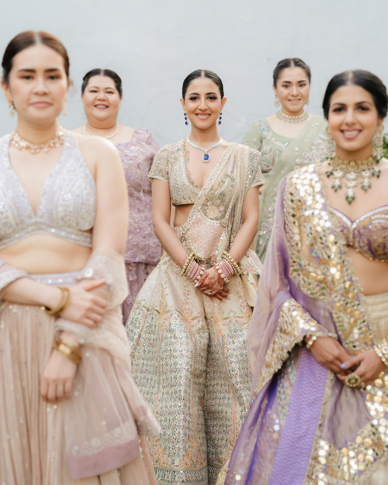 Smiling bride in gold lehenga surrounded by bridesmaids in colorful ethnic wear at Indian wedding.