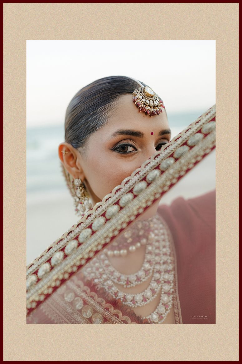 Close-up of Indian bride with maang tikka and pearl-bordered dupatta framing her face at beach wedding.