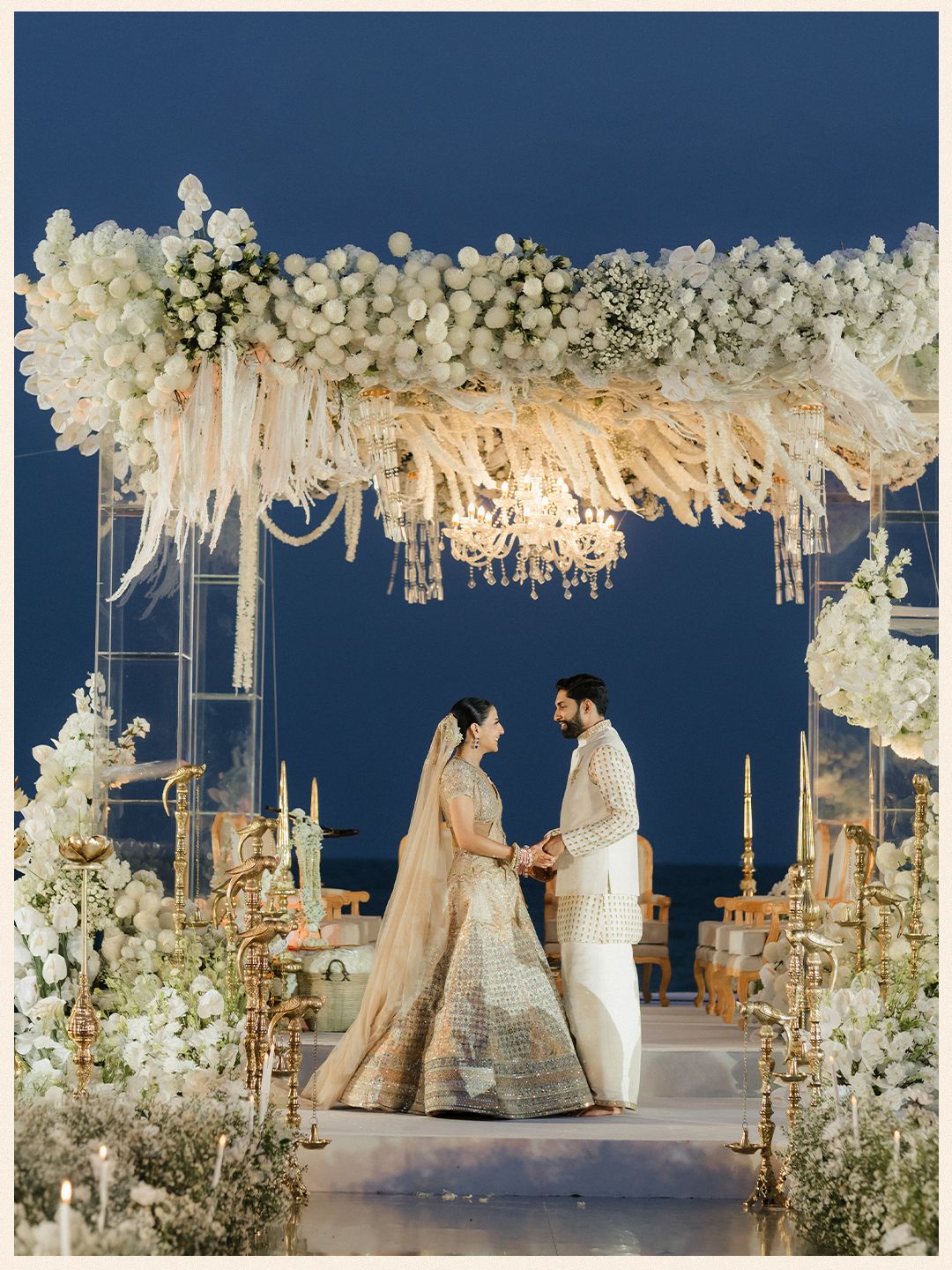 Indian bride and groom exchange vows under a breathtaking white floral mandap at a beach wedding.