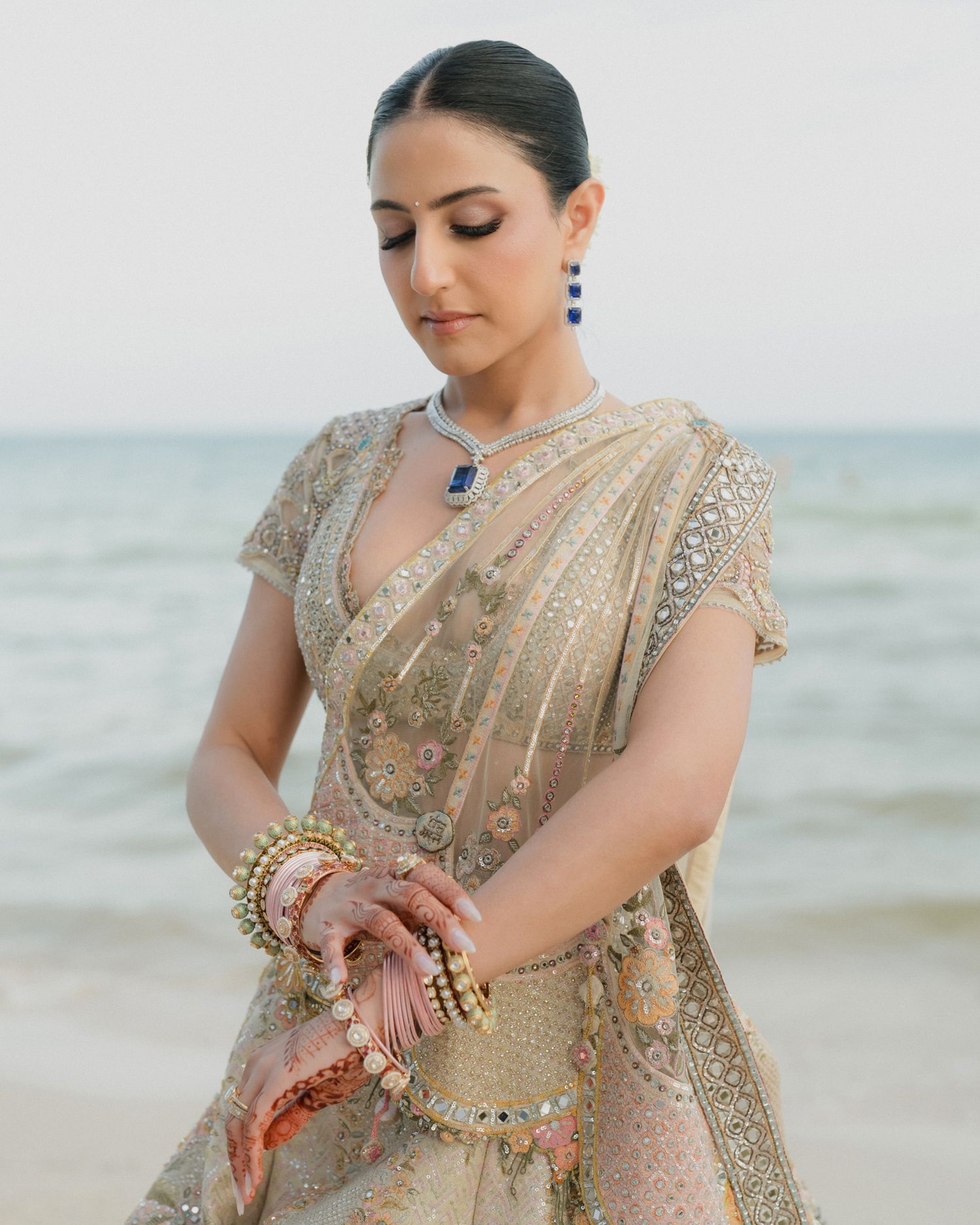 Indian bride in gold embroidered lehenga with sapphire jewelry posing elegantly by the ocean.
