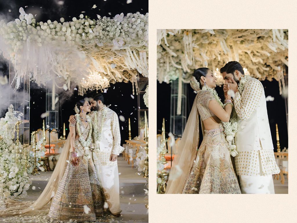 Bride in gold lehenga and groom in white sherwani share romantic moment under white floral arch.