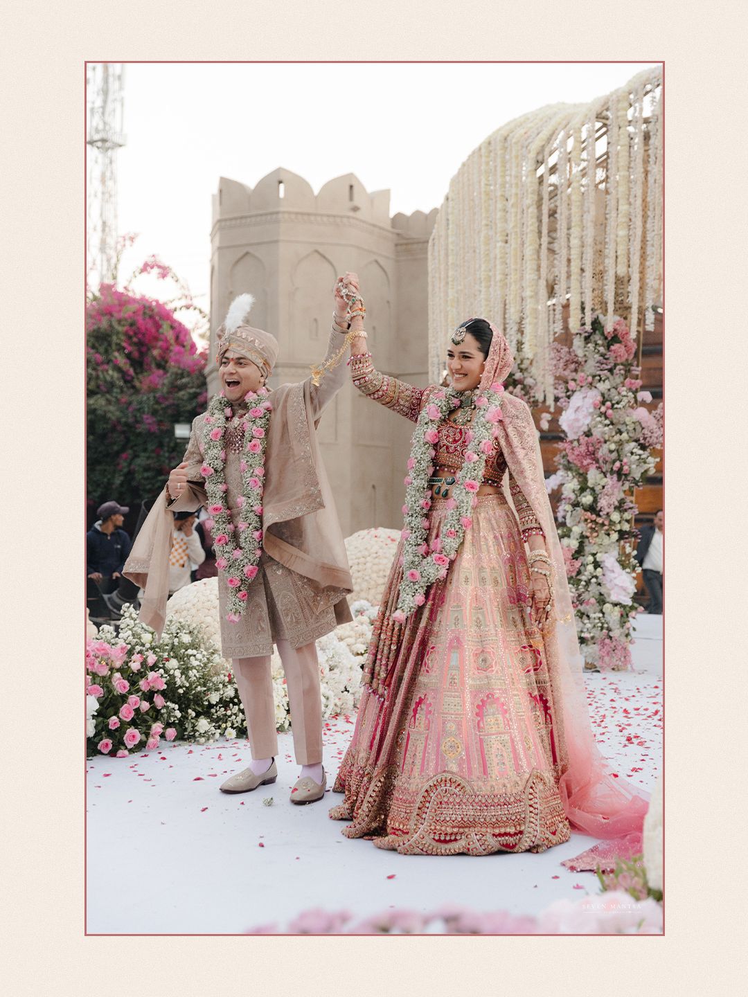 Indian bride in pink lehenga and groom in beige sherwani celebrating joyfully at floral wedding ceremony.