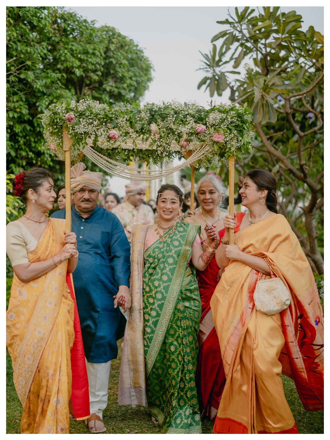 Family members escort bride under floral canopy during traditional Indian wedding procession in lush garden setting.