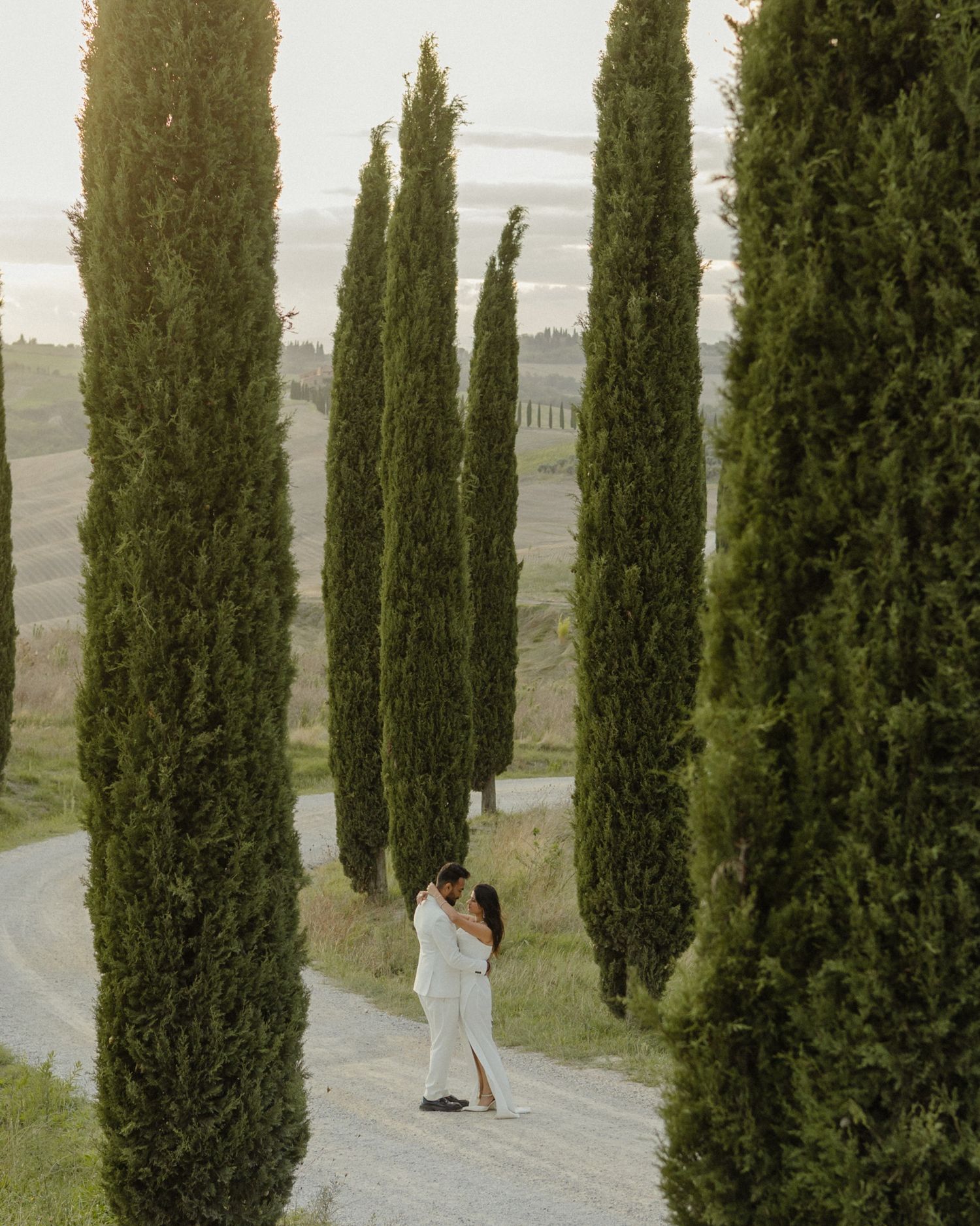 Couple in white outfits embracing on a scenic Tuscan road lined with tall cypress trees at golden hour.