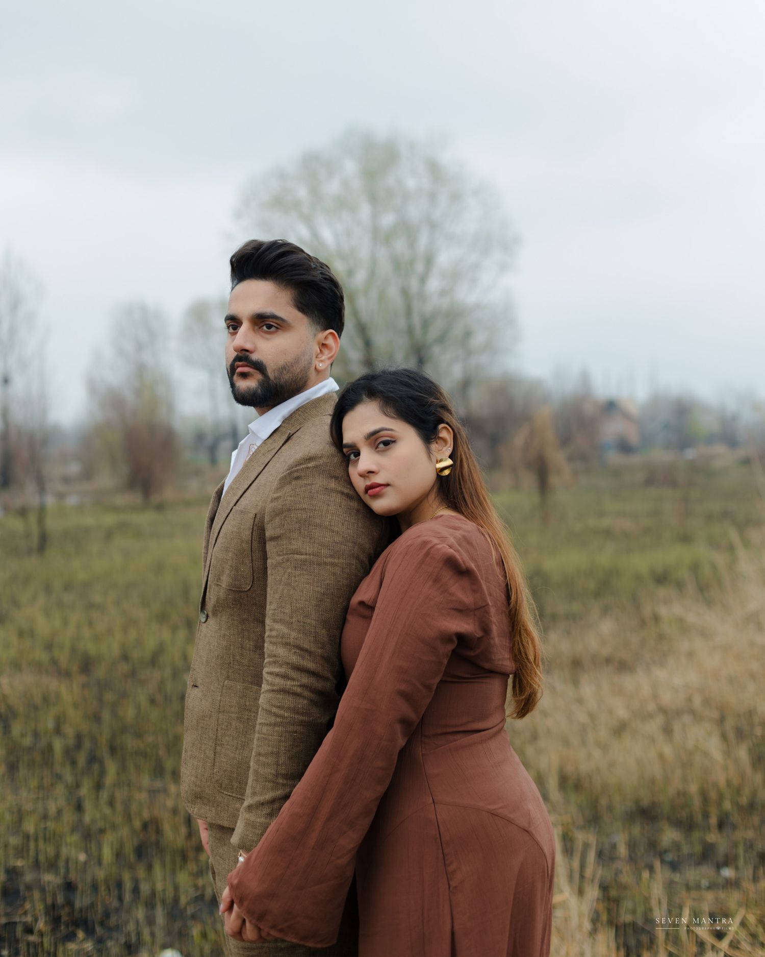 Stylish South Asian couple posing back-to-back in coordinating brown outfits in a moody outdoor field setting.