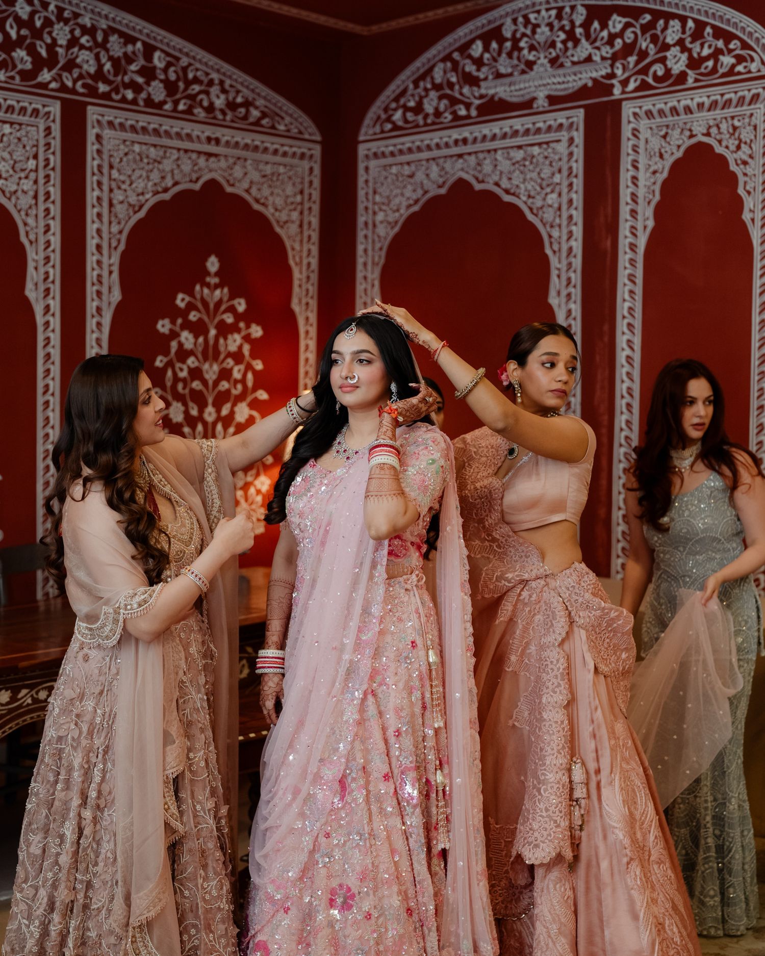Bridesmaids helping bride in pink lehenga prepare in ornate Indian wedding venue.