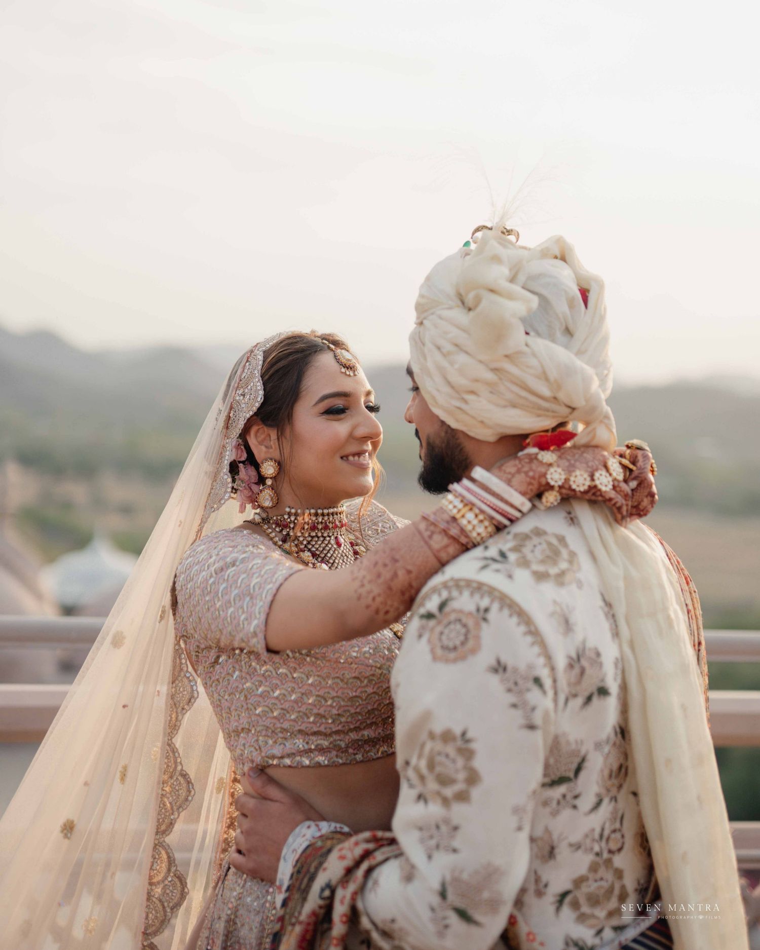 Indian bride and groom sharing joyful moment in traditional wedding attire against scenic backdrop.