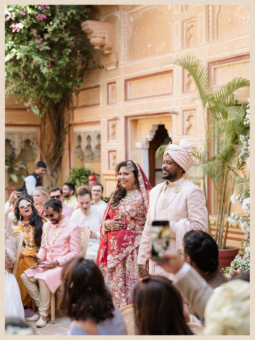 Indian bride and groom walk together during wedding ceremony at ornate Rajasthani palace courtyard with guests seated.