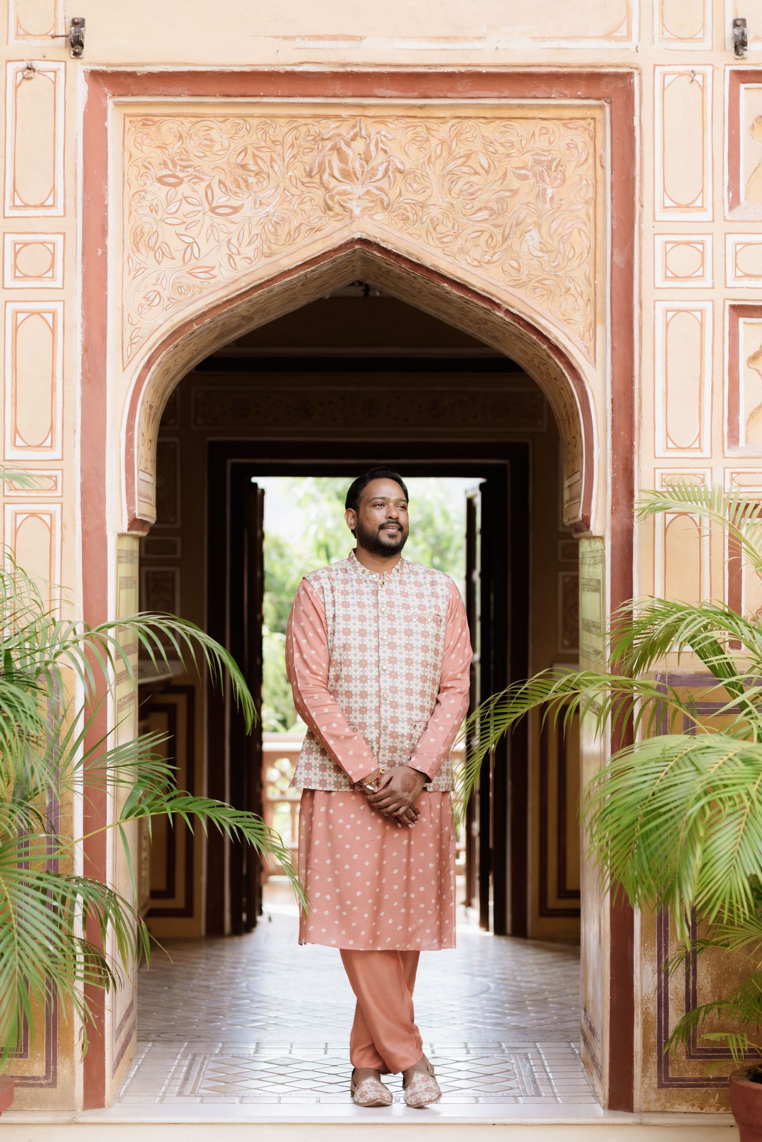 Indian groom in pink kurta standing in ornate Mughal archway with potted palms.