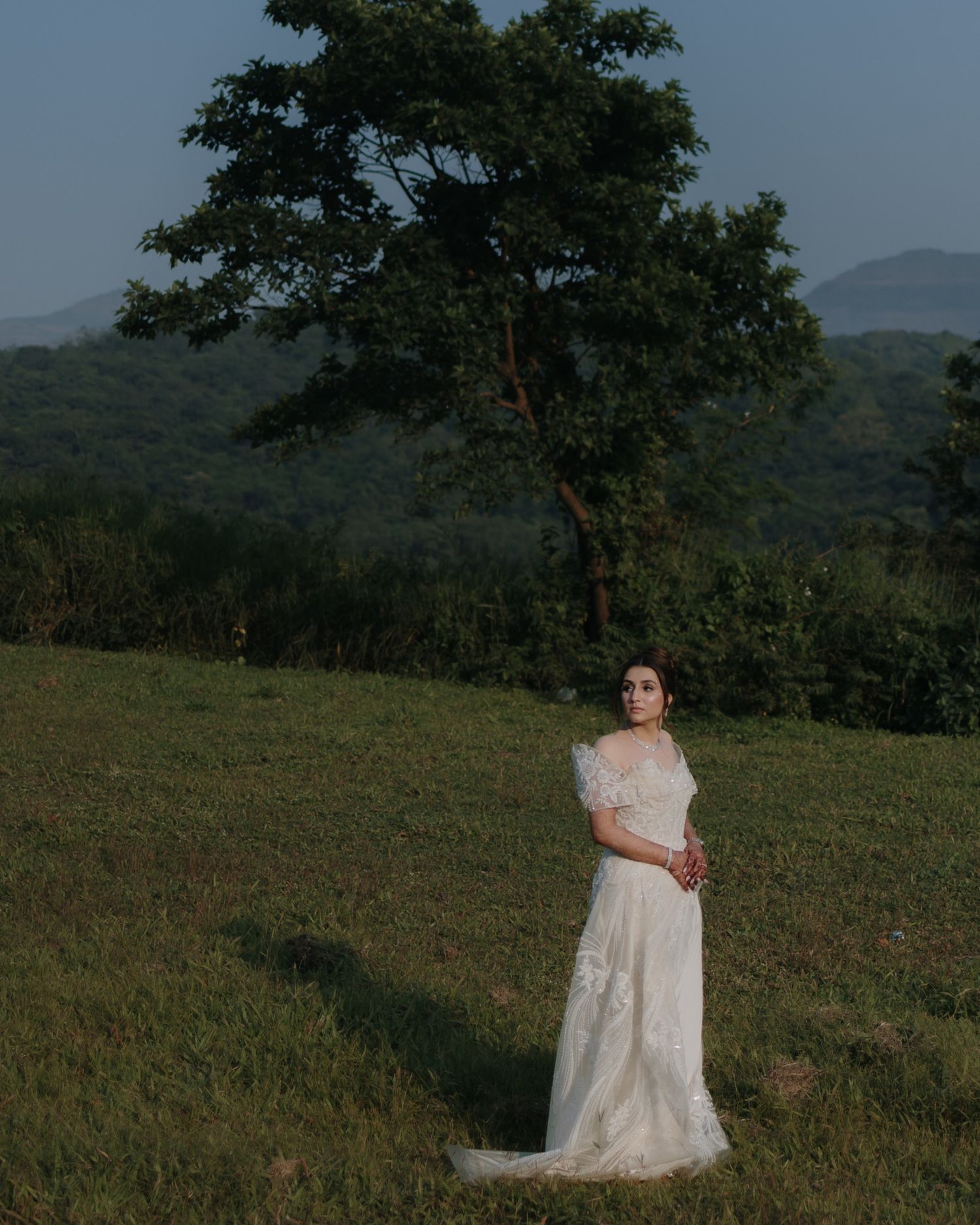 Bride in white lace wedding gown standing in green meadow with large tree and mountain backdrop.