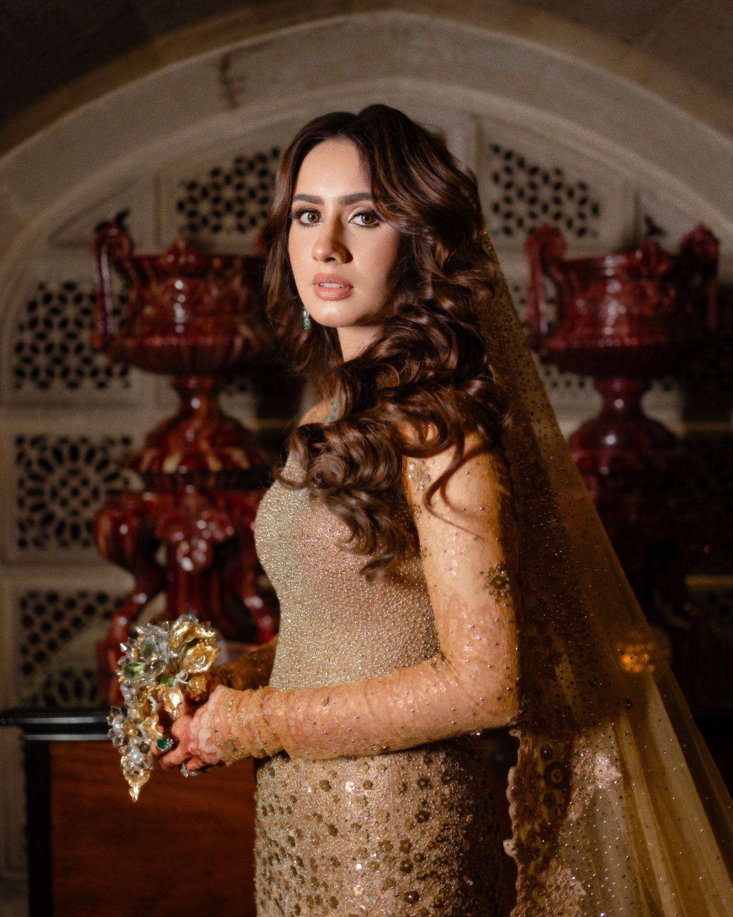 Bride in gold embellished gown with matching veil holding a bouquet in an ornate arched setting.