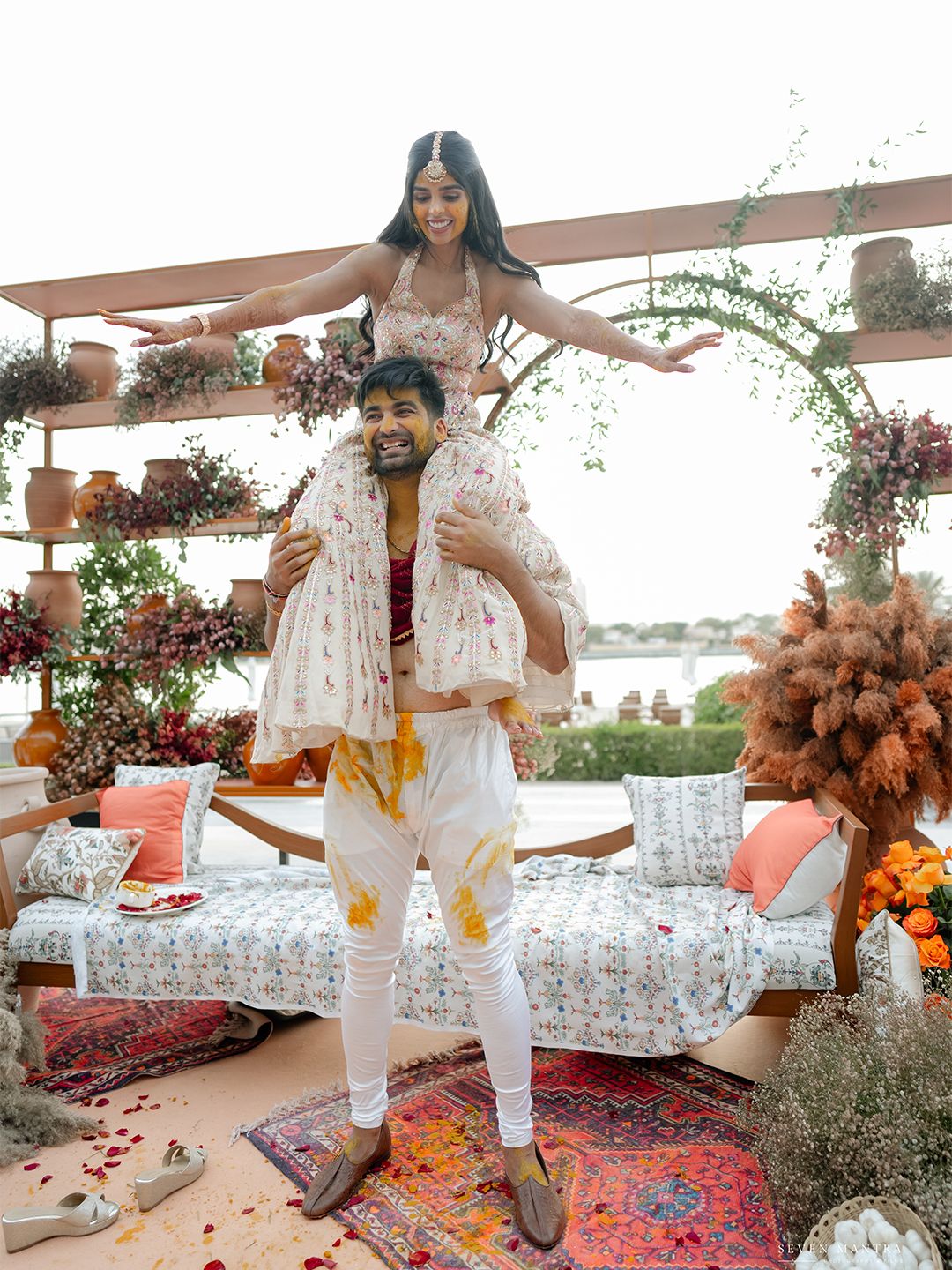 Bride sits on groom's shoulders during Indian Haldi ceremony with floral arch backdrop.
