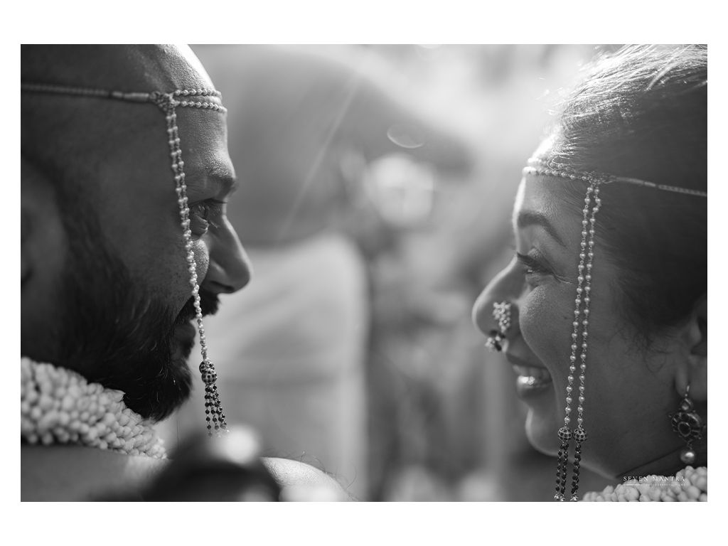 Black and white portrait of Indian bride and groom adorned with maang tikka facing each other.