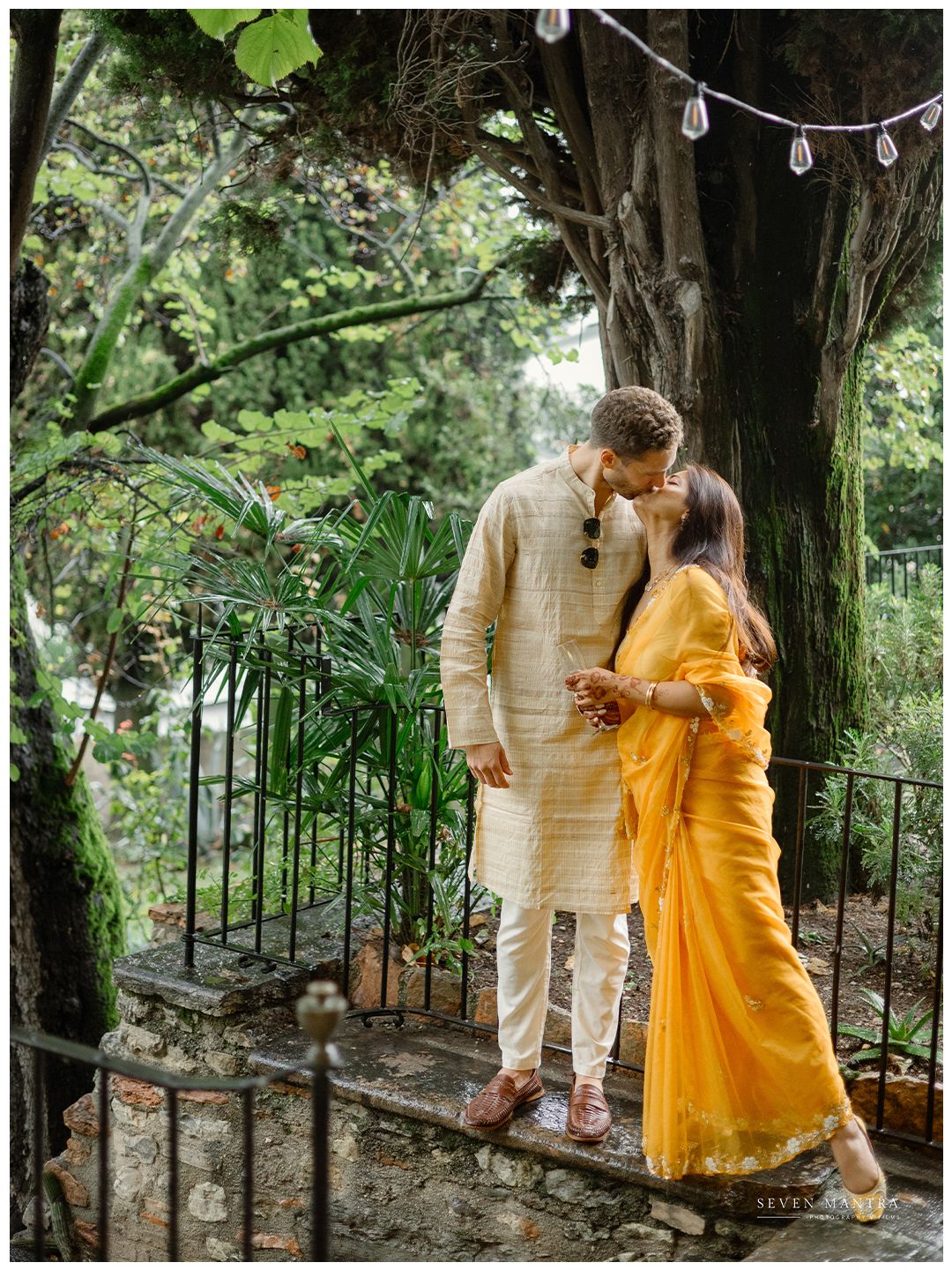 Couple sharing a romantic kiss in a garden, bride wearing a vibrant yellow saree and groom in beige kurta.