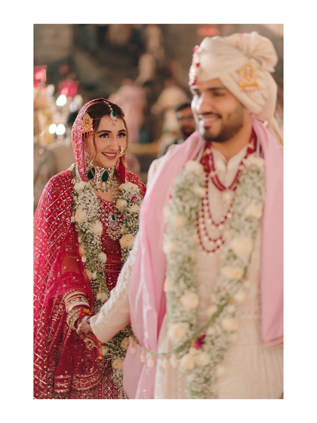 Radiant Indian bride in red lehenga smiling at groom wearing white sherwani during wedding ceremony.