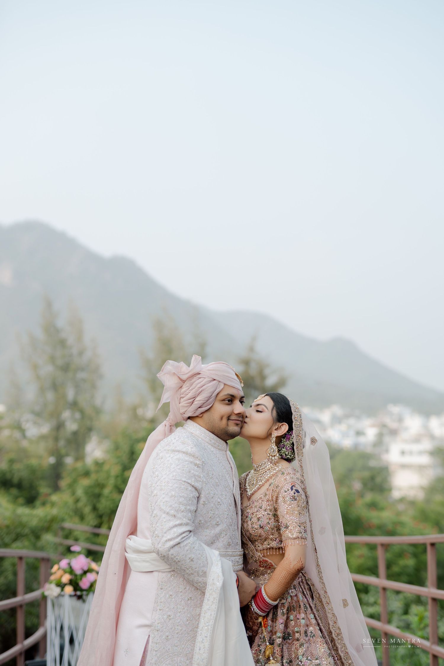 Indian bride kisses groom in white sherwani with pink turban against mountain backdrop at wedding.