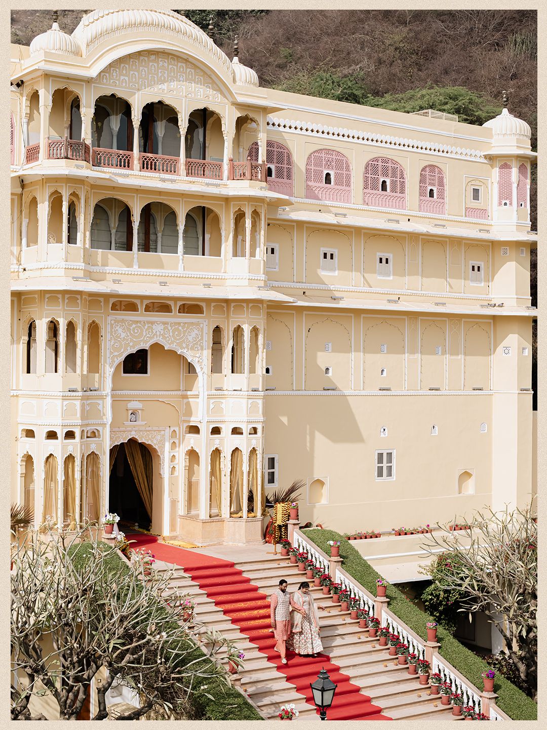 Couple descends grand red carpet staircase of stunning Rajasthani palace wedding venue with ornate pink architecture.