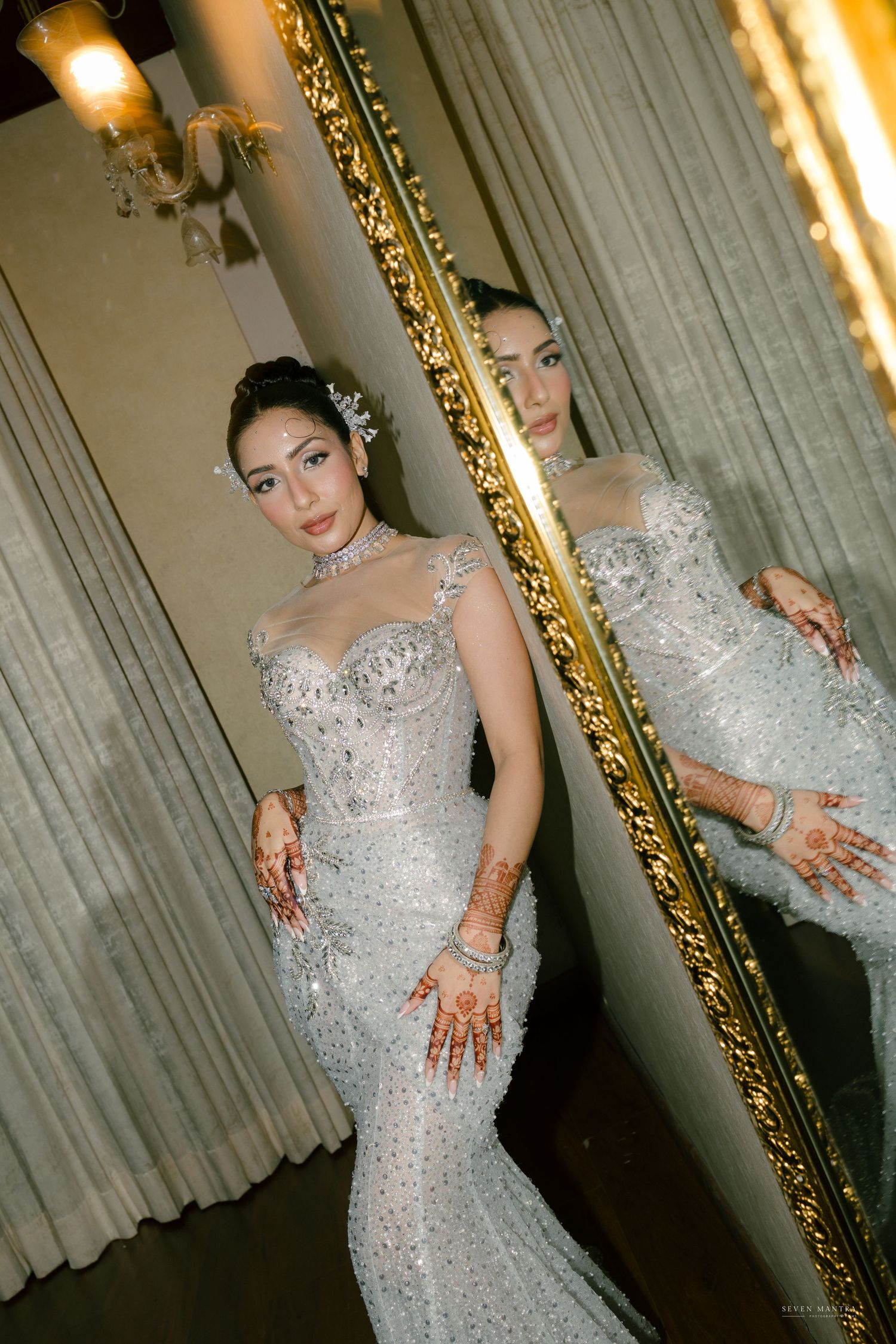 Bride in stunning silver beaded mermaid dress reflected in ornate gold-framed mirror at reception.