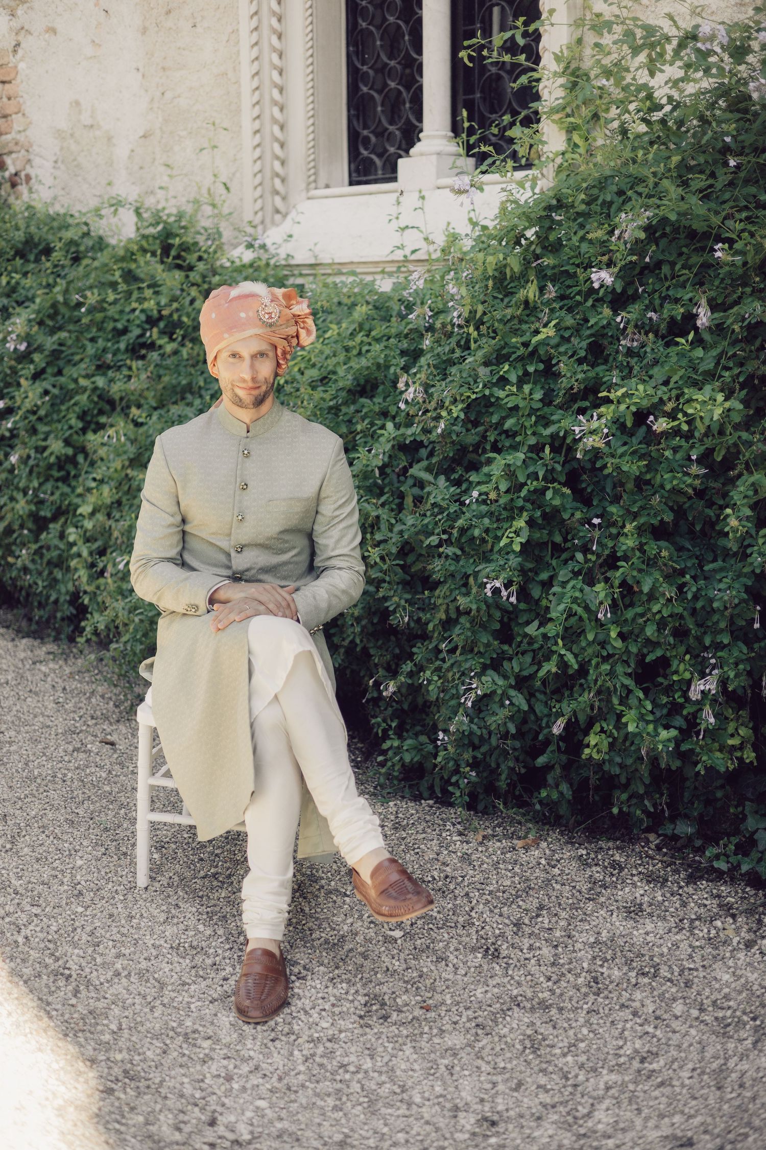 Groom in sage green sherwani and orange turban seated on white chair beside lush garden wall.