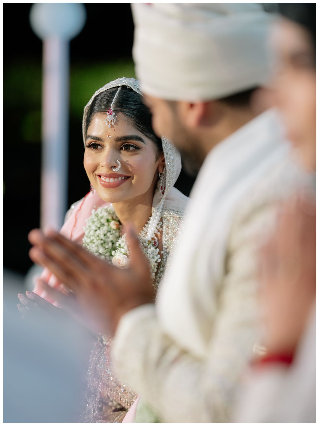 Smiling Indian bride in embroidered lehenga and veil gazes at groom during outdoor wedding ceremony.