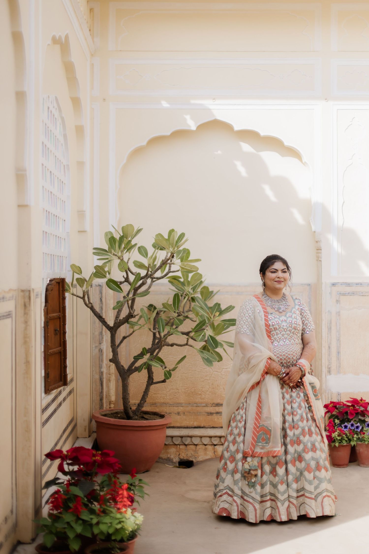 Indian bride in pastel lehenga stands gracefully in a sunlit Rajasthani palace corridor.