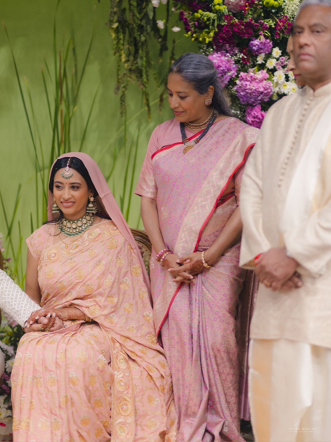 Bride in pink saree sits at wedding ceremony with family members standing beside her.
