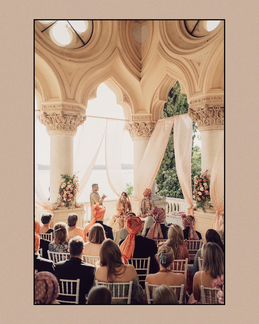 Indian wedding ceremony inside ornate arched venue with floral mandap, guests seated in white chairs.