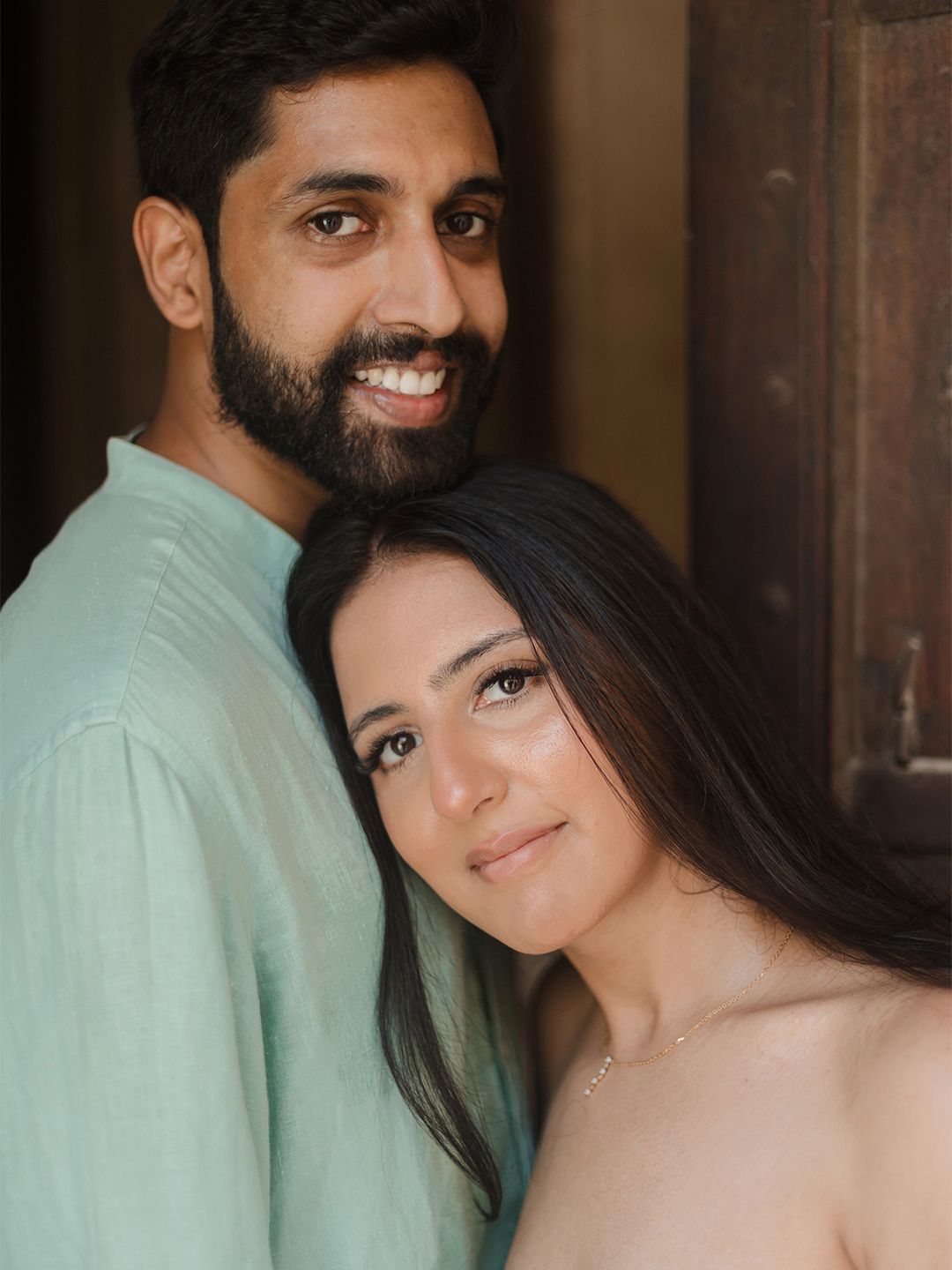 Smiling Indian couple in casual mint kurta and nude attire posing closely together in front of wooden door.
