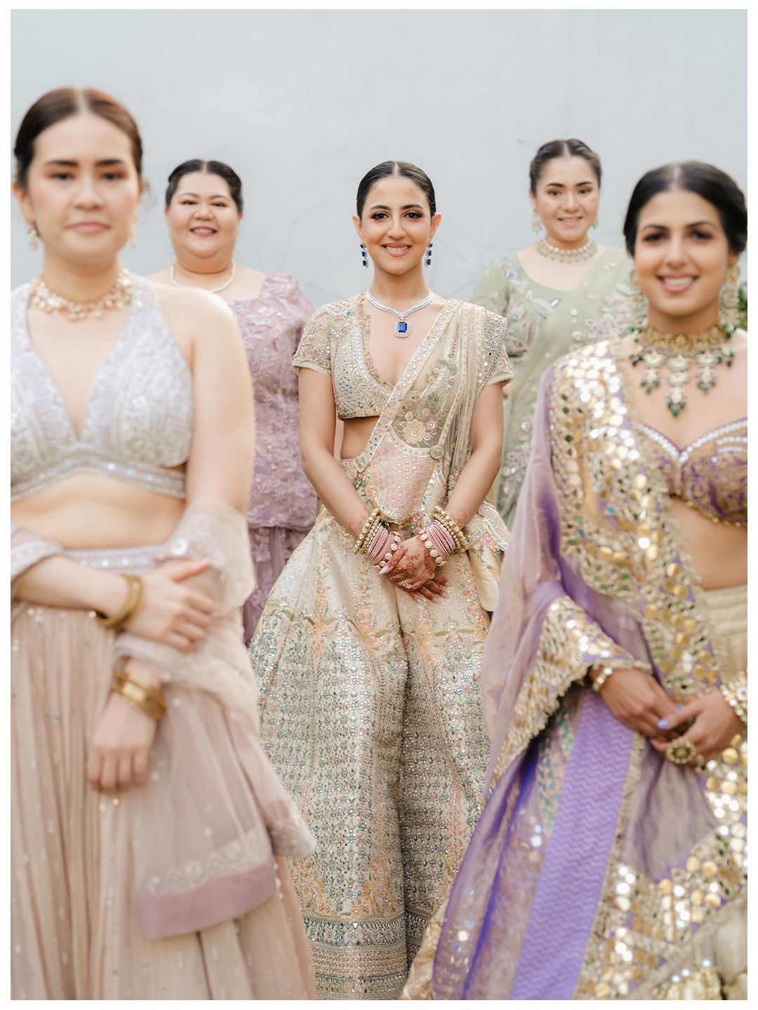 Indian bride in gold lehenga with sapphire necklace poses with bridesmaids in pastel outfits at wedding.