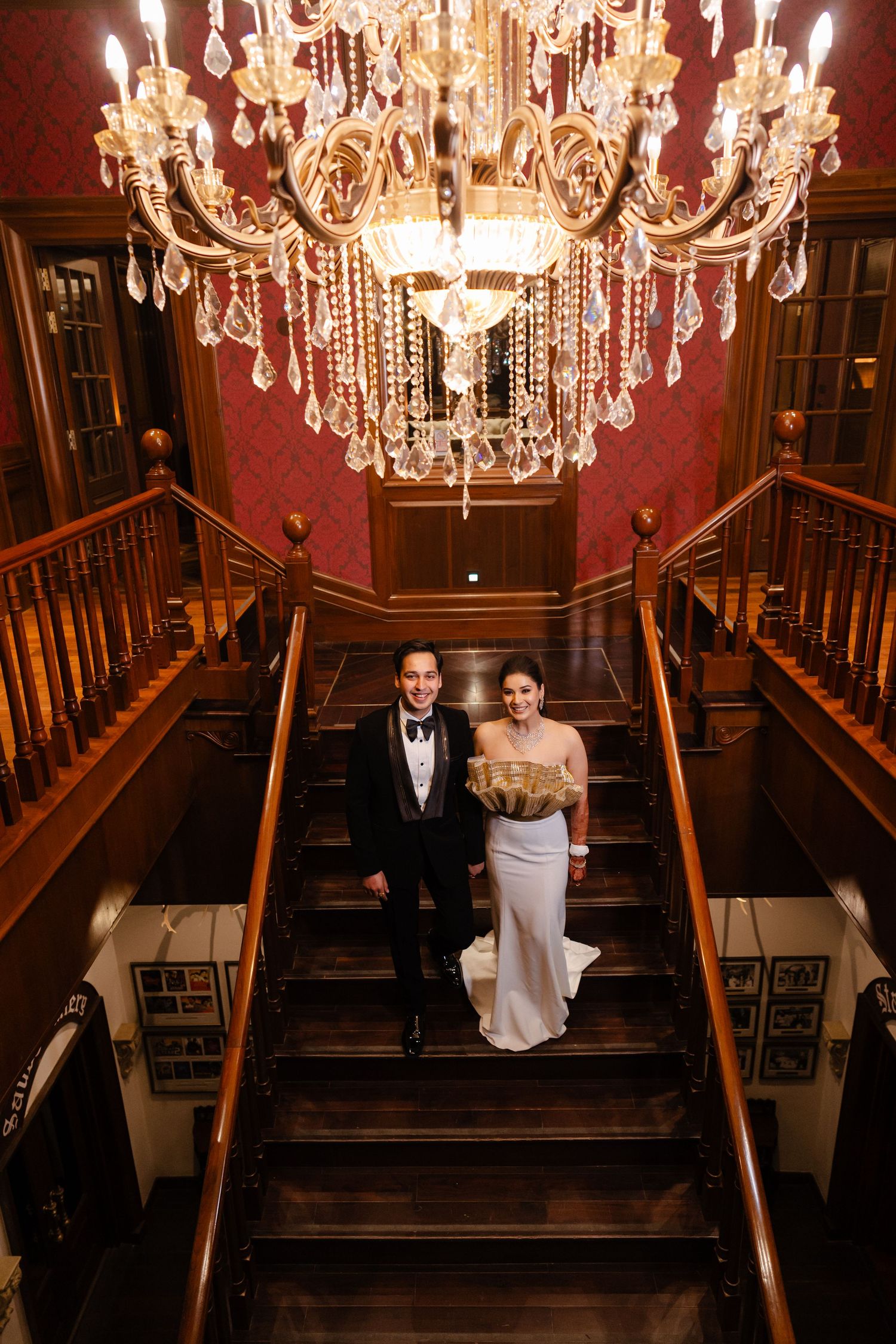 Bride and groom standing on grand staircase beneath ornate crystal chandelier in luxurious wedding venue.