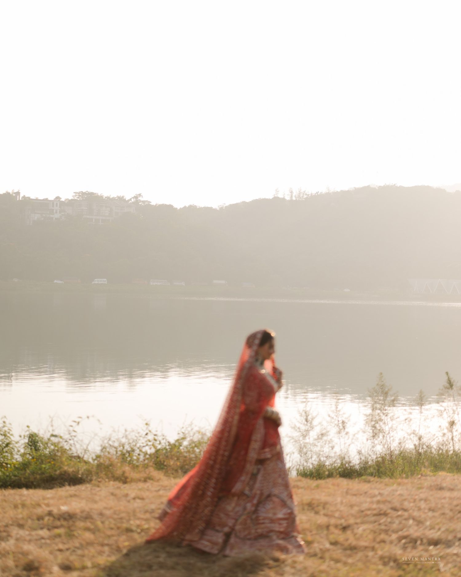 Indian bride in red lehenga with dupatta flowing in breeze beside serene misty lake at dawn.