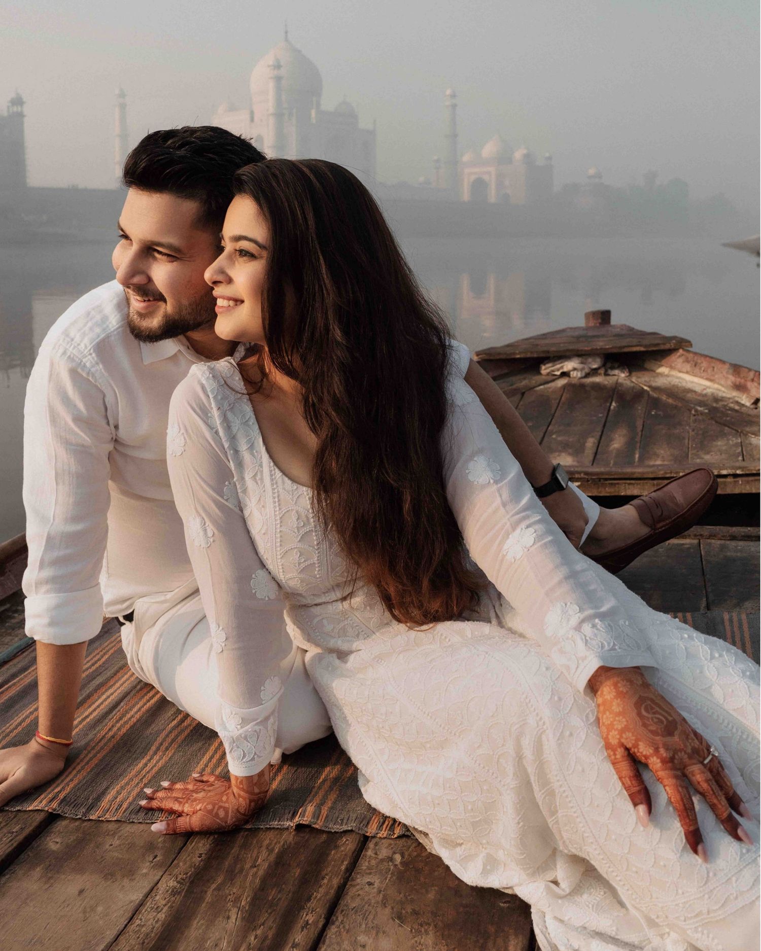 Romantic couple in white outfits sitting on wooden dock with Taj Mahal backdrop at sunrise.