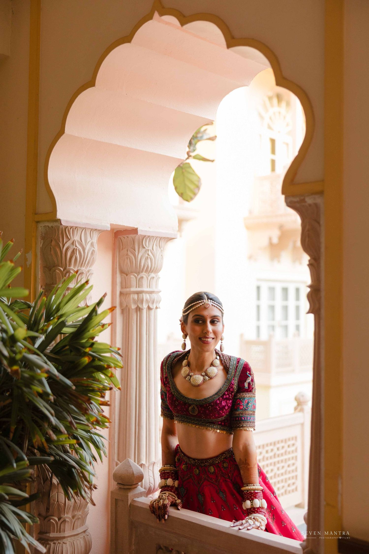 Smiling bride in red lehenga adorned with traditional jewelry standing in carved white palace archway.
