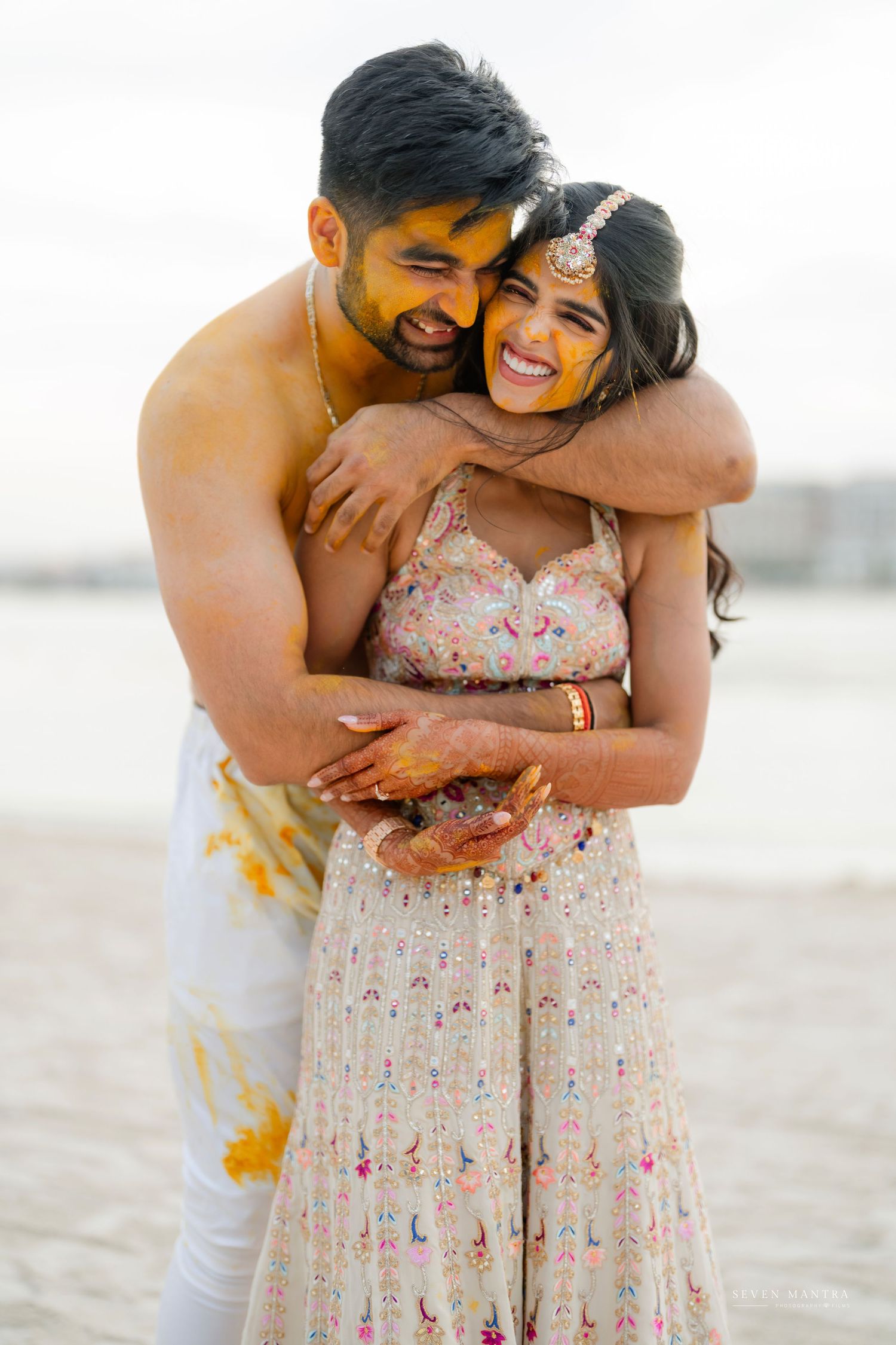 Couple laughing joyfully covered in haldi turmeric during beach haldi ceremony pre-wedding celebration.
