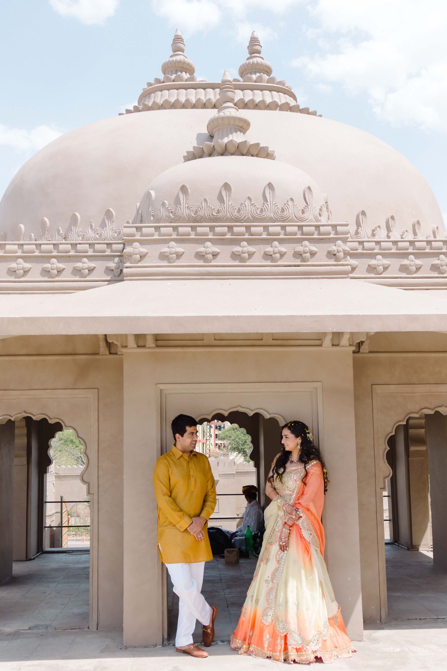 Indian couple in traditional attire standing under ornate temple dome during pre-wedding photoshoot.