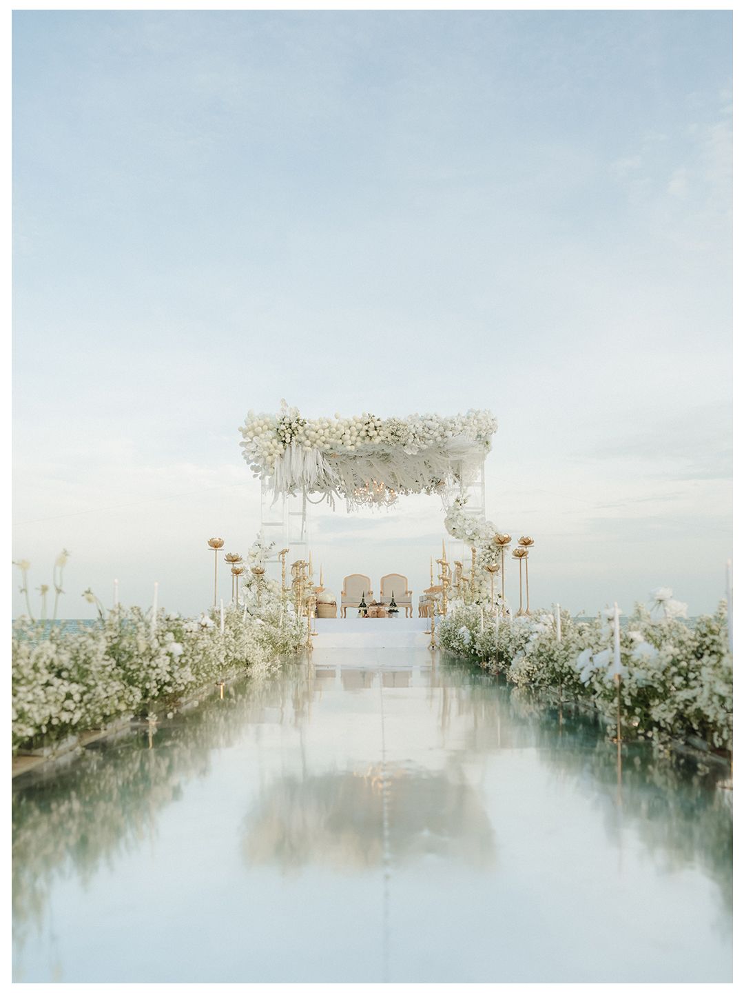 Elegant white floral wedding ceremony arch with mirrored aisle and golden accents overlooking ocean at sunset.