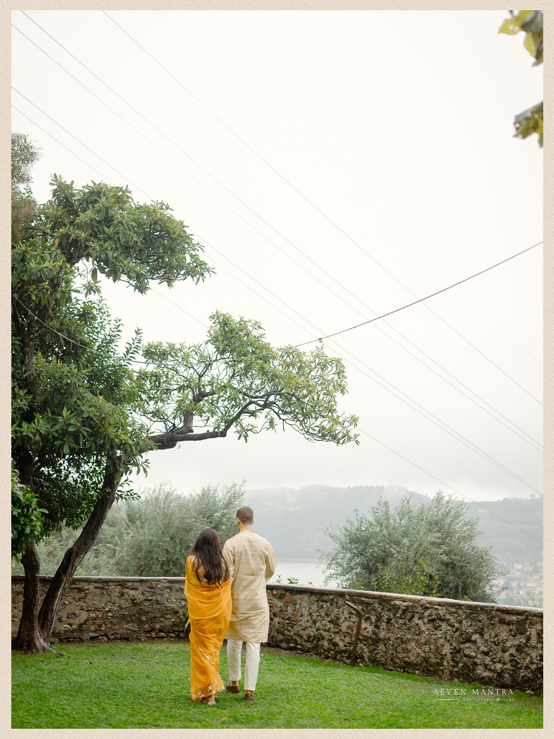 Bride in yellow saree and groom in beige kurta standing together overlooking a misty Italian landscape.