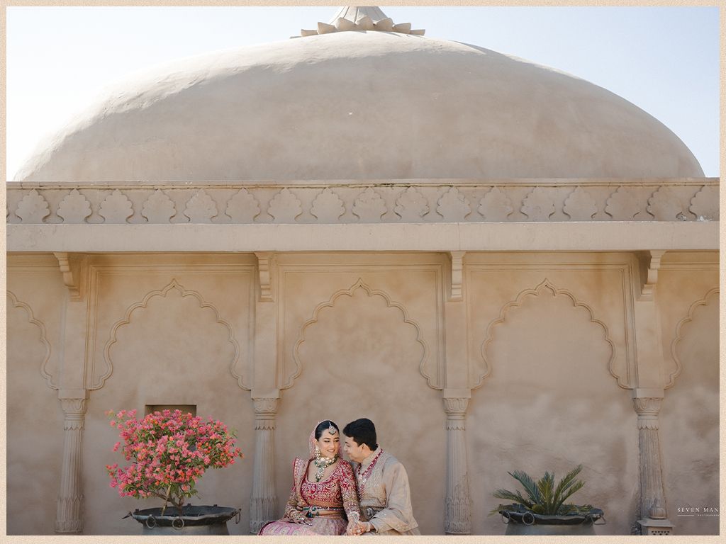 Bride and groom sit together beneath a white dome with pink bougainvillea at Jaipur palace.