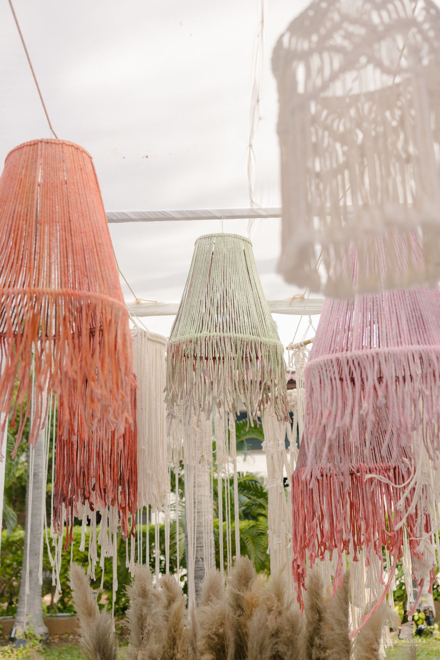 Colorful boho macrame fringe chandeliers in pink, coral and white hanging outdoors at a wedding decor display.