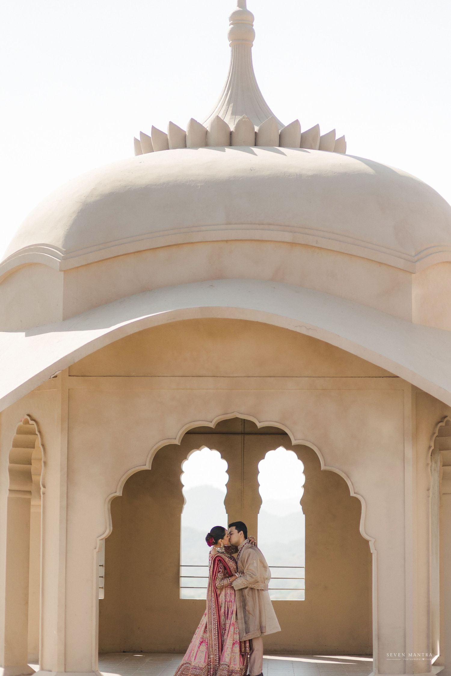 Indian bride and groom share intimate moment under white marble arch at Jaipur palace wedding.