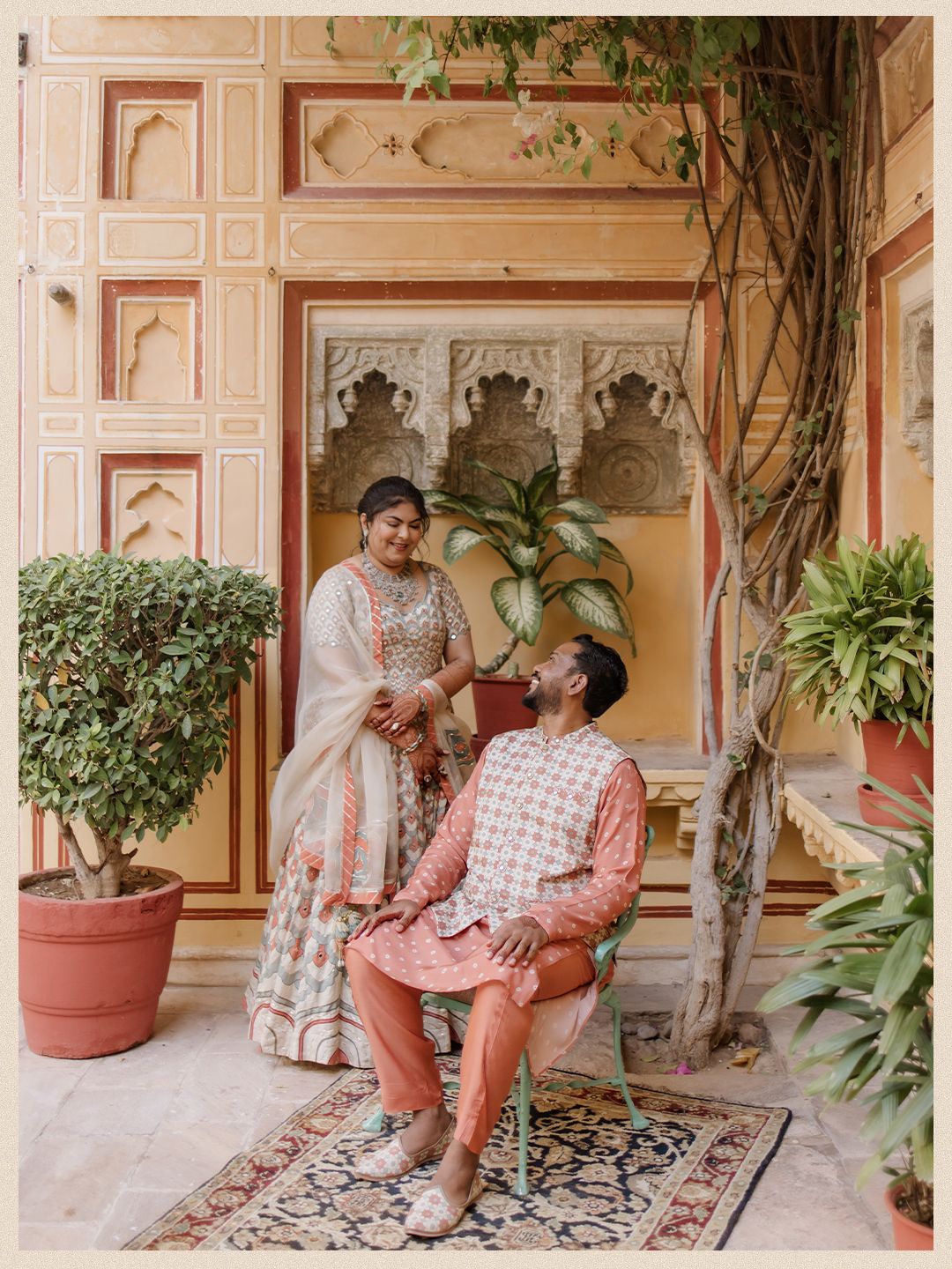 Indian bride and groom share romantic moment in ornate Rajasthani courtyard surrounded by lush tropical plants.