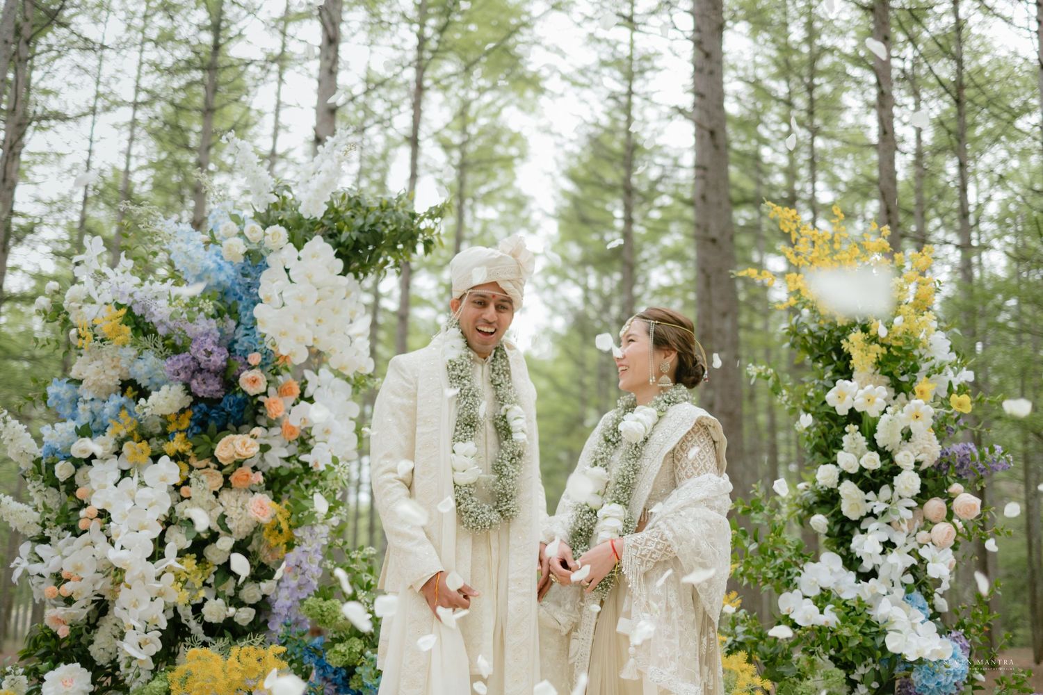 Couple in white traditional Indian attire holding hands under floral arch in a serene forest wedding.