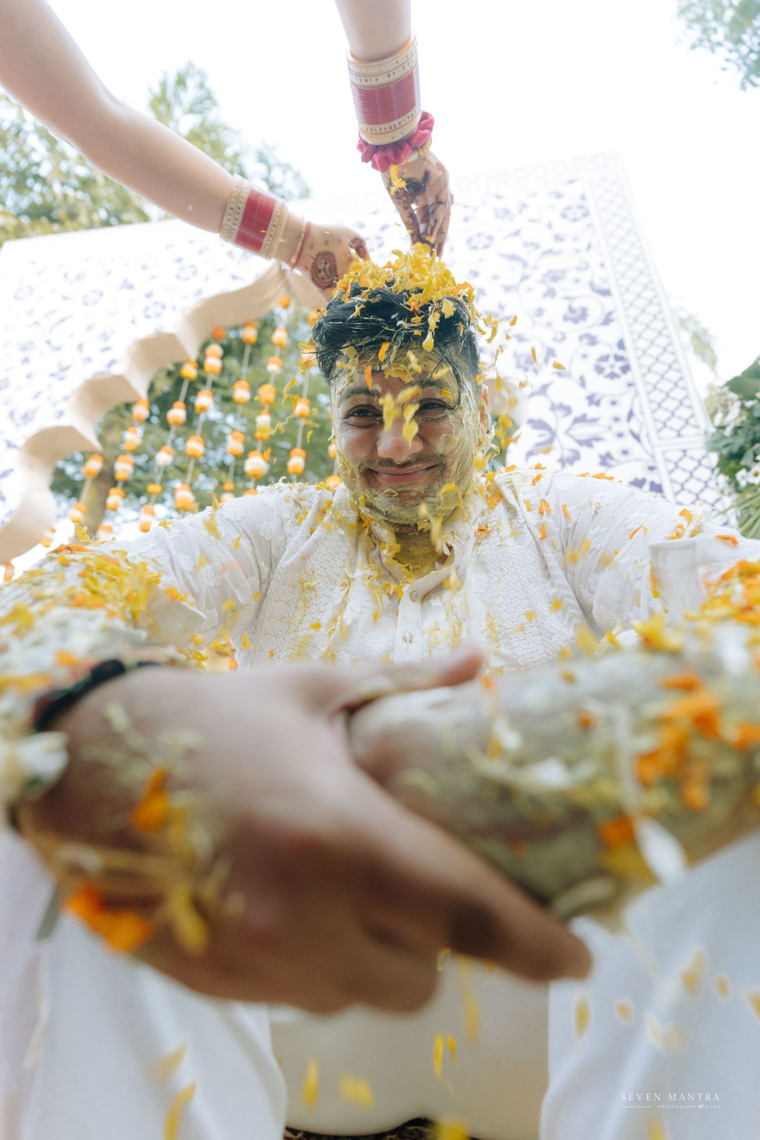 Groom laughing joyfully during haldi ceremony as turmeric paste and marigold flowers are applied.
