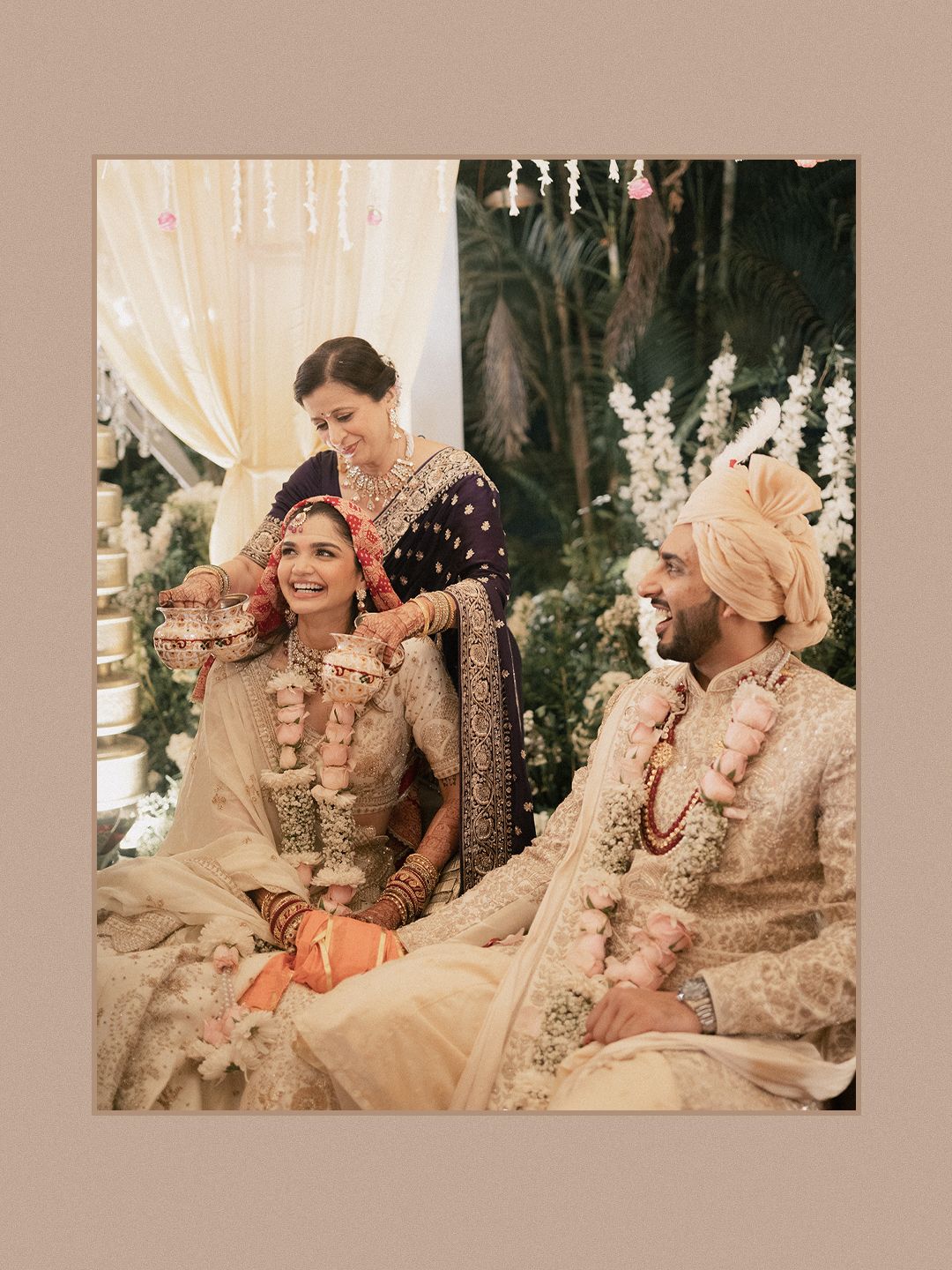 Bride and groom in cream embroidered outfits seated at floral Indian wedding ceremony mandap.