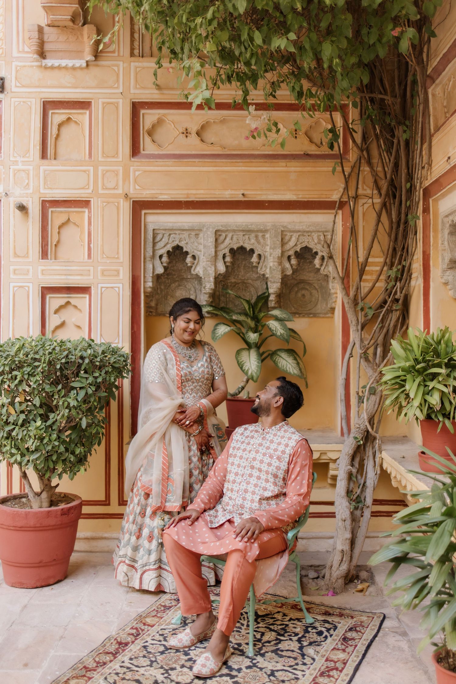 Indian couple in coordinating pink outfits share a romantic moment in an ornate palace courtyard.