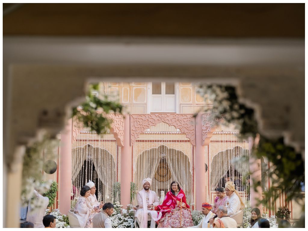 Indian wedding ceremony captured through archway at a grand pink Rajasthani palace with guests and priest seated.
