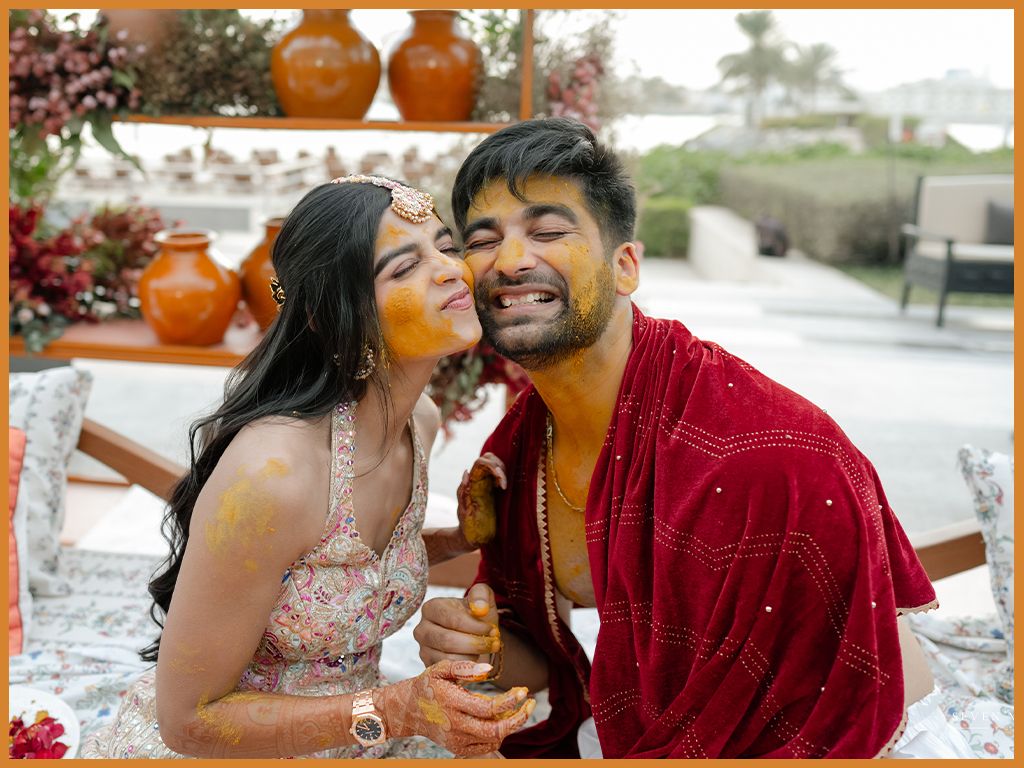 Bride kisses groom's cheek during Haldi ceremony, both covered in turmeric paste.
